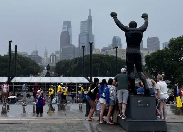 Tourists pose with the Rocky statue on the steps of the Philadelphia Museum of Art on Saturday, June 27, 2026. In the background, the concert stage for the city's July 4 concert can be seen being built by crews ahead of the show.
